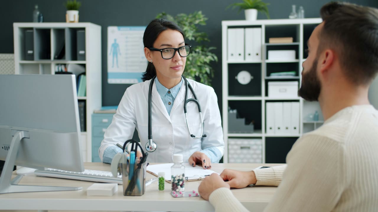 Doctor in a white coat talking with a patient across a desk in a clinic consultation room